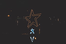 Neon signs shaped like stars and letters are illuminated against a dark background. The larger star glows in yellow, while a smaller one shines in blue beneath it.