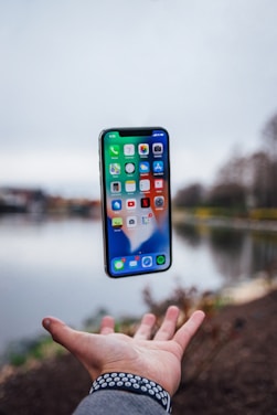 A smartphone appears to be levitating above an outstretched hand with a blurred scenic background of a lake and trees. The screen displays various app icons, indicating it is turned on. The hand is adorned with a patterned bracelet and sleeve of a jacket.