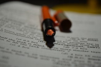 A close-up of a fountain pen with an orange cap and barrel lies on a sheet of paper filled with printed text and mathematical problems. The focus is on the nib of the pen, while the text in the background is blurred.
