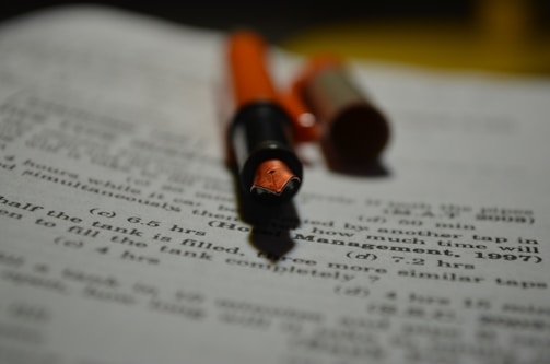 A close-up of a fountain pen with an orange cap and barrel lies on a sheet of paper filled with printed text and mathematical problems. The focus is on the nib of the pen, while the text in the background is blurred.