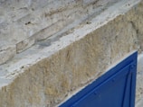 A textured stone wall with visible layers, featuring a pronounced horizontal groove, displaying shades of beige and tan. In the foreground, a bright blue metal structure or panel offers a contrast to the natural stone hues.