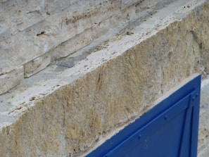 A textured stone wall with visible layers, featuring a pronounced horizontal groove, displaying shades of beige and tan. In the foreground, a bright blue metal structure or panel offers a contrast to the natural stone hues.