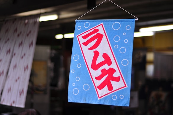 A blue fabric sign featuring red Japanese characters centrally displayed against a backdrop of white bubbles. The sign is hanging in a slightly blurred indoor setting, possibly a shop or market.