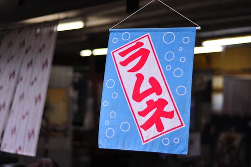 A blue fabric sign featuring red Japanese characters centrally displayed against a backdrop of white bubbles. The sign is hanging in a slightly blurred indoor setting, possibly a shop or market.