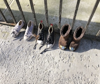 A set of four shoes is lined up against a wrought iron railing. Two pairs of shoes include white sneakers and black sneakers with white toe caps, as well as brown fleece-lined boots. The ground is a textured, light gray surface, possibly concrete, with shadows cast by the railing.