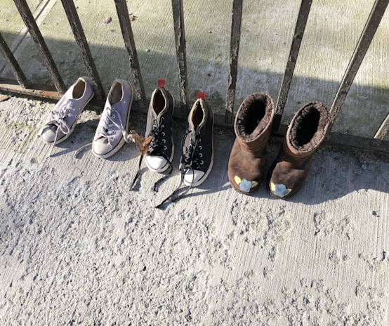 A set of four shoes is lined up against a wrought iron railing. Two pairs of shoes include white sneakers and black sneakers with white toe caps, as well as brown fleece-lined boots. The ground is a textured, light gray surface, possibly concrete, with shadows cast by the railing.