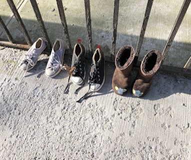 A set of four shoes is lined up against a wrought iron railing. Two pairs of shoes include white sneakers and black sneakers with white toe caps, as well as brown fleece-lined boots. The ground is a textured, light gray surface, possibly concrete, with shadows cast by the railing.