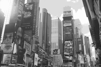 A bustling urban scene with tall buildings adorned with large billboards and advertisements. The view focuses on a crowded area featuring multiple colorful signs promoting various brands and events. Skyscrapers rise in the background, creating a dense cityscape.