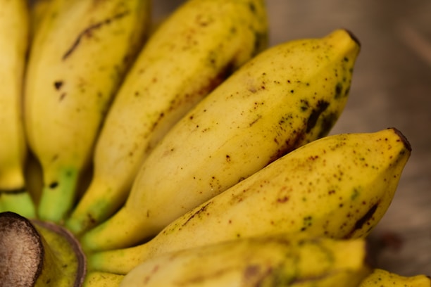 A close-up of ripe bananas with a smooth yellow skin and some brown spots, arranged together on a wooden surface. The bananas appear fresh and ready to eat.