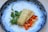 A gourmet dish featuring a piece of grilled or baked fish, garnished with herbs and small green leafy vegetables, accompanied by a colorful mix of diced carrots and possibly another root vegetable. The plate itself has a distinct design with a speckled blue rim.