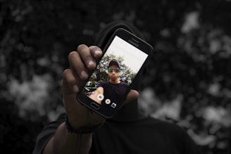 A person holds a Samsung smartphone displaying a selfie in focus. The background is blurred, highlighting the phone and its screen displaying a person wearing sunglasses and a cap.