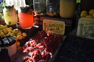 An outdoor stall features large glass jars filled with colorful beverages, including pink and yellow lemonade, accompanied by freshly cut lemons used as garnishes. Fresh lemons, strawberries, and blueberries are displayed prominently alongside. The setup is accented by rustic wooden crates and vintage-style signs advertising ice-cold lemonade.