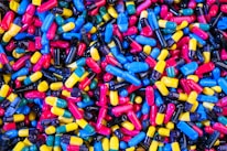 A large collection of colorful capsules and pills, including blue, yellow, red, black, and multi-colored combinations, scattered in a random pattern.