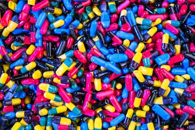 A large collection of colorful capsules and pills, including blue, yellow, red, black, and multi-colored combinations, scattered in a random pattern.