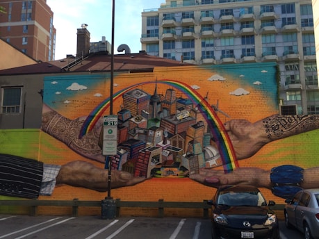 A vibrant mural depicting a pair of hands holding a colorful miniature cityscape with skyscrapers and a bridge. A rainbow arches over the city, set against a bright, gradient sky with clouds. The hands have intricate designs and tattoos on them.