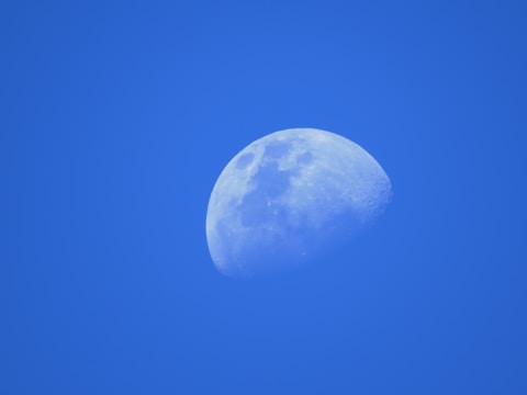 A clear half-moon is set against a bright blue sky, showcasing detailed lunar surface features.