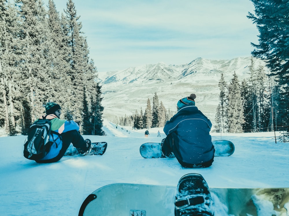 two persons sitting on snow with their snowboards