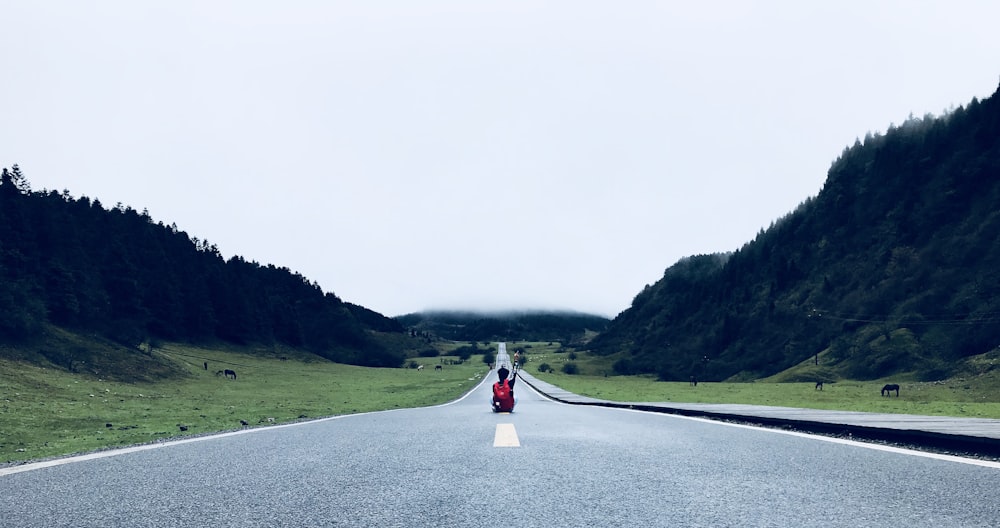 person sitting on gray road in between mountains under white sky at daytime