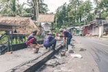 Several workers are engaged in construction or repair work along a roadside. They are using various tools and materials, such as buckets and spades. The environment includes a rural road with palm trees and traditional thatched-roof buildings, including a sign for a cafe.