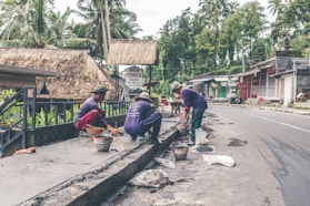Several workers are engaged in construction or repair work along a roadside. They are using various tools and materials, such as buckets and spades. The environment includes a rural road with palm trees and traditional thatched-roof buildings, including a sign for a cafe.