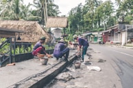 Several workers are engaged in construction or repair work along a roadside. They are using various tools and materials, such as buckets and spades. The environment includes a rural road with palm trees and traditional thatched-roof buildings, including a sign for a cafe.