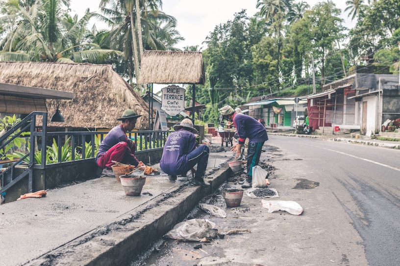 Several workers are engaged in construction or repair work along a roadside. They are using various tools and materials, such as buckets and spades. The environment includes a rural road with palm trees and traditional thatched-roof buildings, including a sign for a cafe.