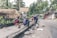 Several workers are engaged in construction or repair work along a roadside. They are using various tools and materials, such as buckets and spades. The environment includes a rural road with palm trees and traditional thatched-roof buildings, including a sign for a cafe.