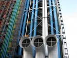 An architectural facade featuring a series of colorful pipes and ducts, including blue, green, and red vertical conduits. Large white cylindrical vents with grid-like openings dominate the foreground. The structure displays exposed industrial elements with numerous steel beams and cables.