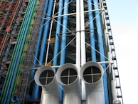 An architectural facade featuring a series of colorful pipes and ducts, including blue, green, and red vertical conduits. Large white cylindrical vents with grid-like openings dominate the foreground. The structure displays exposed industrial elements with numerous steel beams and cables.