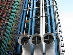 An architectural facade featuring a series of colorful pipes and ducts, including blue, green, and red vertical conduits. Large white cylindrical vents with grid-like openings dominate the foreground. The structure displays exposed industrial elements with numerous steel beams and cables.