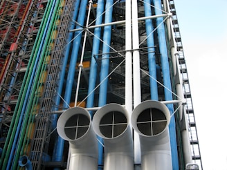 An architectural facade featuring a series of colorful pipes and ducts, including blue, green, and red vertical conduits. Large white cylindrical vents with grid-like openings dominate the foreground. The structure displays exposed industrial elements with numerous steel beams and cables.