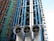 An architectural facade featuring a series of colorful pipes and ducts, including blue, green, and red vertical conduits. Large white cylindrical vents with grid-like openings dominate the foreground. The structure displays exposed industrial elements with numerous steel beams and cables.