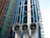 An architectural facade featuring a series of colorful pipes and ducts, including blue, green, and red vertical conduits. Large white cylindrical vents with grid-like openings dominate the foreground. The structure displays exposed industrial elements with numerous steel beams and cables.