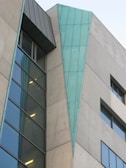A modern building facade features a combination of concrete and large glass panels. A distinctive triangular teal-blue metallic element runs vertically along part of the structure, adding a unique architectural detail. Windows of varying sizes are present, some reflecting light, contributing to a sleek and contemporary design.