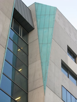 A modern building facade features a combination of concrete and large glass panels. A distinctive triangular teal-blue metallic element runs vertically along part of the structure, adding a unique architectural detail. Windows of varying sizes are present, some reflecting light, contributing to a sleek and contemporary design.