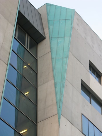 A modern building facade features a combination of concrete and large glass panels. A distinctive triangular teal-blue metallic element runs vertically along part of the structure, adding a unique architectural detail. Windows of varying sizes are present, some reflecting light, contributing to a sleek and contemporary design.