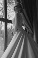 A woman in an elegant, long bridal gown stands next to a large window, with natural light highlighting the intricate lace design of the dress. Her hair is styled in an elaborate updo, adorned with decorative accessories. The background shows bare tree branches through the window, indicating a scene set in winter or late fall.