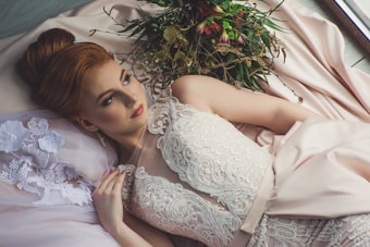 A woman lies elegantly in a luxurious setting, wearing a lace-embellished gown. Her hair is styled in an updo, and she gazes thoughtfully into the distance. A bouquet of dried flowers is placed beside her, and the fabric drapes softly around her, adding to the serene ambiance.