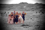 A group of people, including a bride in a white gown, are walking barefoot on a beach. The bridesmaids are wearing dark blue dresses and holding bouquets of yellow and red flowers. The background features expansive sand dunes in grayscale.