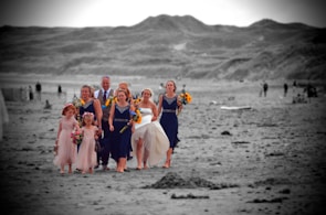 A group of people, including a bride in a white gown, are walking barefoot on a beach. The bridesmaids are wearing dark blue dresses and holding bouquets of yellow and red flowers. The background features expansive sand dunes in grayscale.