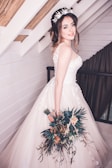 A bride is standing indoors, wearing a beautiful white wedding gown with floral lace details. She has a bouquet of flowers in her left hand and is wearing a floral headband. The background features white wooden walls with exposed beams and a curtain.