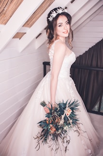 A bride is standing indoors, wearing a beautiful white wedding gown with floral lace details. She has a bouquet of flowers in her left hand and is wearing a floral headband. The background features white wooden walls with exposed beams and a curtain.