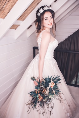 A bride is standing indoors, wearing a beautiful white wedding gown with floral lace details. She has a bouquet of flowers in her left hand and is wearing a floral headband. The background features white wooden walls with exposed beams and a curtain.