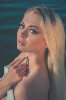 A woman with long blond hair is posing with a gentle expression and has her hand near her chin. Her eyes are highlighted with makeup, and the background is a dark blue water surface.