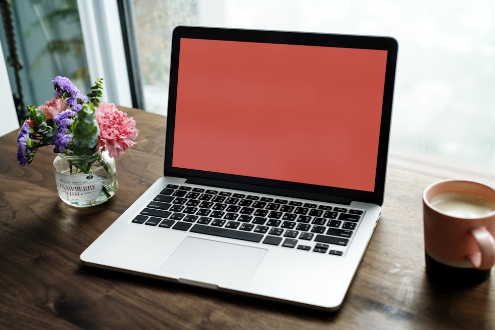 MacBook Pro on wooden surface between mug and flower arrangement