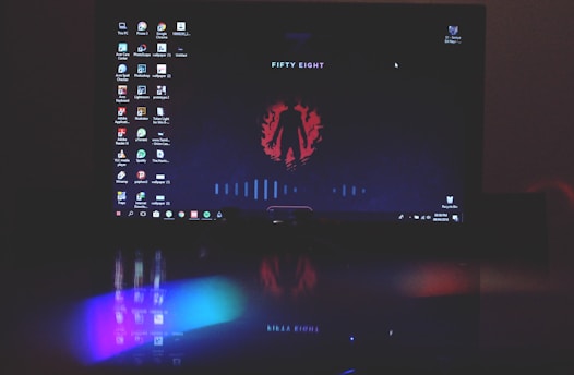 A computer screen displays a dark-themed desktop background with an abstract red emblem and the words 'FIFTY EIGHT'. Various desktop icons are aligned along the left side. The lower part of the image includes a reflection showing vibrant, colorful lights, possibly from a keyboard or other LED device below the screen.