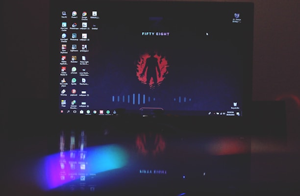 A computer screen displays a dark-themed desktop background with an abstract red emblem and the words 'FIFTY EIGHT'. Various desktop icons are aligned along the left side. The lower part of the image includes a reflection showing vibrant, colorful lights, possibly from a keyboard or other LED device below the screen.