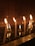 Several white candles are lit, casting a warm glow against a dark, reflective surface that appears to be water. The background features a tiled wall that absorbs the light, creating a serene and contemplative atmosphere. Some wax has dripped and congealed on the surface below, enhancing the sense of tranquility.