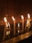 Several white candles are lit, casting a warm glow against a dark, reflective surface that appears to be water. The background features a tiled wall that absorbs the light, creating a serene and contemplative atmosphere. Some wax has dripped and congealed on the surface below, enhancing the sense of tranquility.