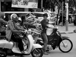 Two people are riding motorbikes on a busy street. The individual on the left wears a helmet and a jacket, while the passenger on the right appears to be carrying a large bundle of clothes on hangers, balanced on the motorcycle. There are cars and tuk-tuks in the background, indicating an urban setting.